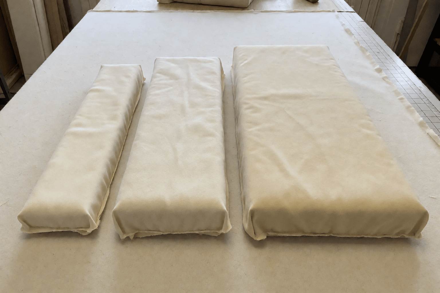 Three cream and neutral fabric-wrapped foam pads of different widths and depths lying in a neat row on an upholsterer's cutting table